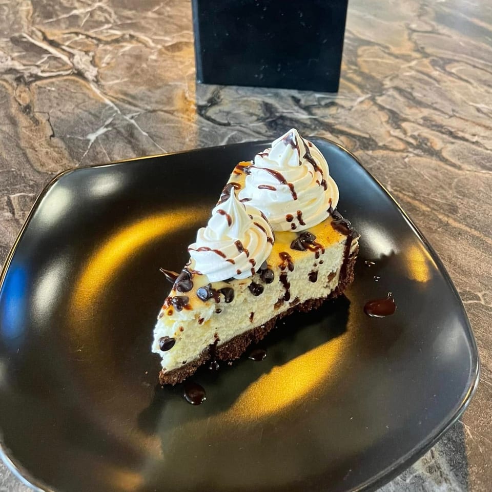 A slice of cheesecake with chocolate chips, whipped cream, and chocolate drizzle on a black plate.