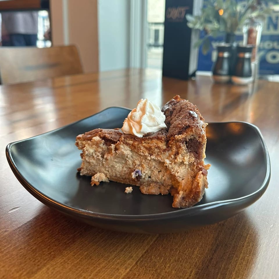 A slice of cheesecake with whipped cream on a black plate at Crust Pizzeria.