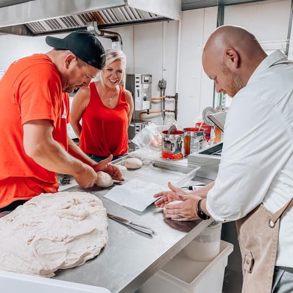 Three people in a commercial kitchen preparing pizza dough on a stainless steel counter.