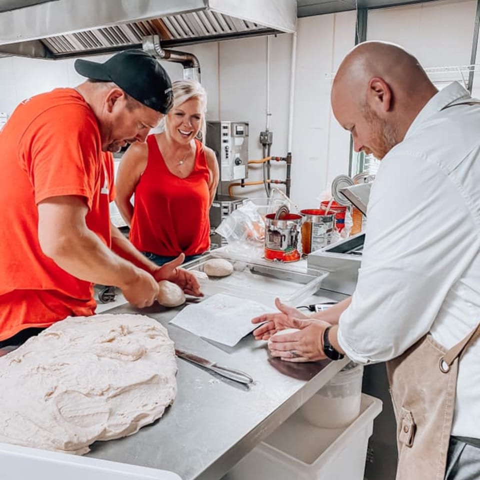 Three people in a commercial kitchen preparing pizza dough on a stainless steel counter.