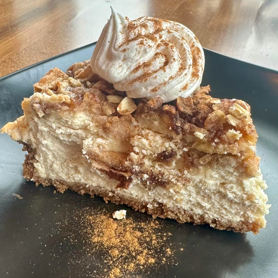 A slice of cheesecake topped with whipped cream, cinnamon, and oat crumble on a black plate.