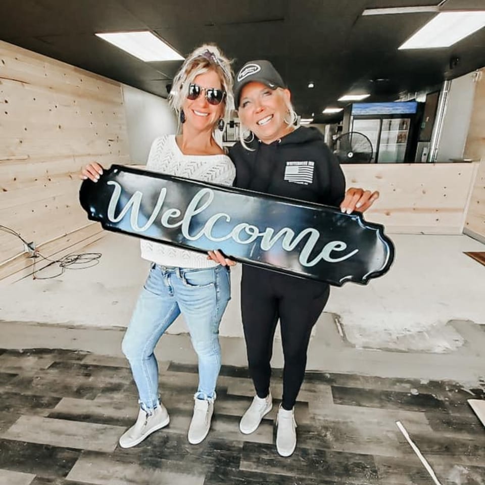 Two smiling women holding a "Welcome" sign inside the Crust Pizzeria restaurant under construction.
