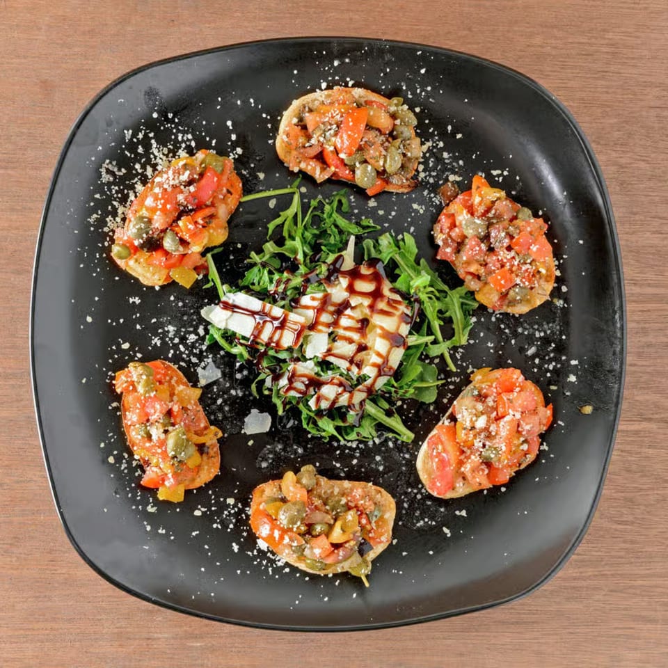 Six bruschetta pieces with tomato, capers, and cheese, surrounding a bed of arugula with cheese.