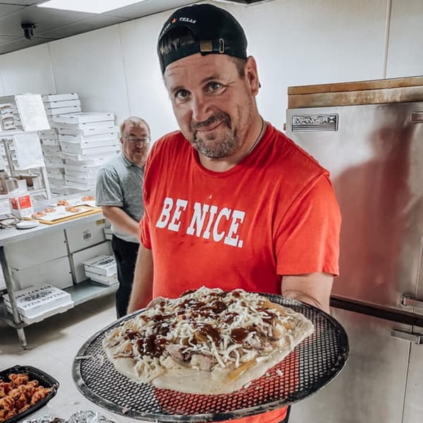 A man in a red "BE NICE" shirt holds an uncooked BBQ chicken pizza at Crust Pizzeria.